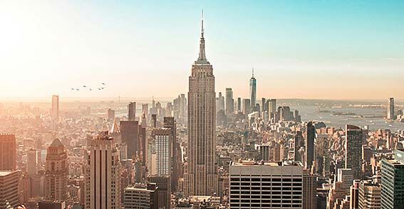 The New York skyline as seen from the south, with the Empire State Building at its centre against a light blue sky.